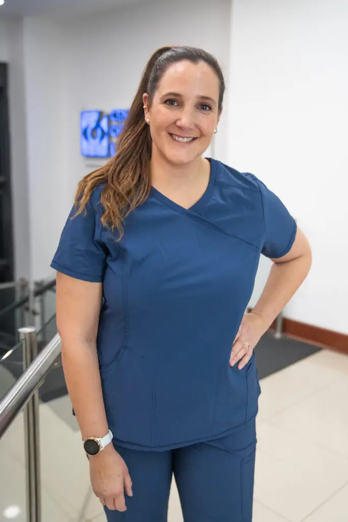 A smiling woman with a ponytail wearing blue scrubs in a medical office.